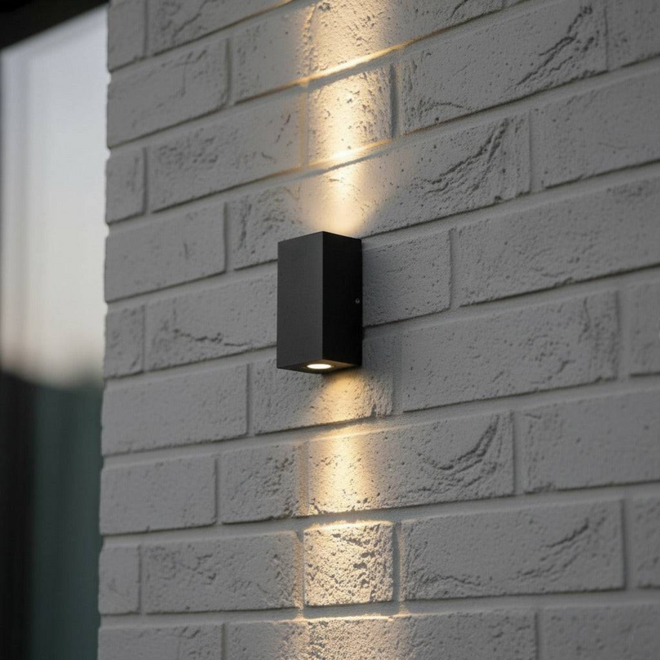 Ultra Beam Lighting Ltd 6W black aluminium square LED wall light casting dual-direction beams on a grey brick wall.