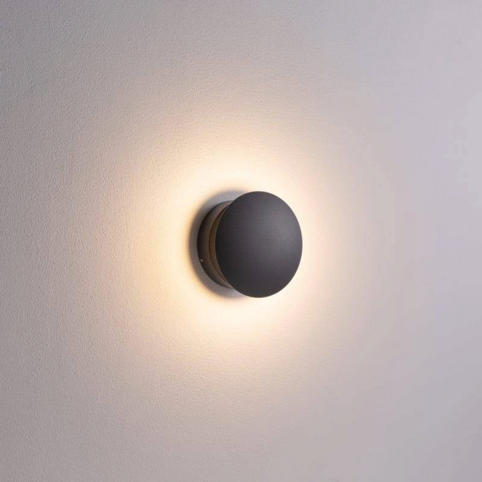 Ultra Beam Lighting Ltd 6W round black LED wall light fixture casting a warm ambient glow on a plain interior wall.
